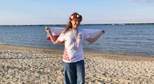 Na plaży stoi dziewczyna z medalem na szyi, wiankiem w barwach narodowych i flagą biało-czerwoną.