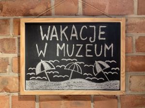 Na ceglanej ścianie powieszona tablica z napisem kredą: wakacje w muzeum oraz parasolkami wbitymi w piasek.