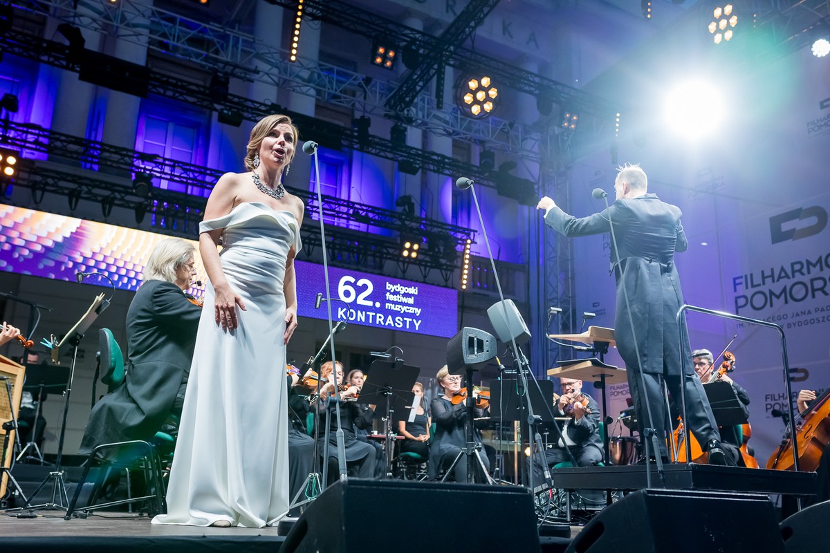 Śpiewaczka w białej sukni na scenie z orkiestrą symfoniczną.