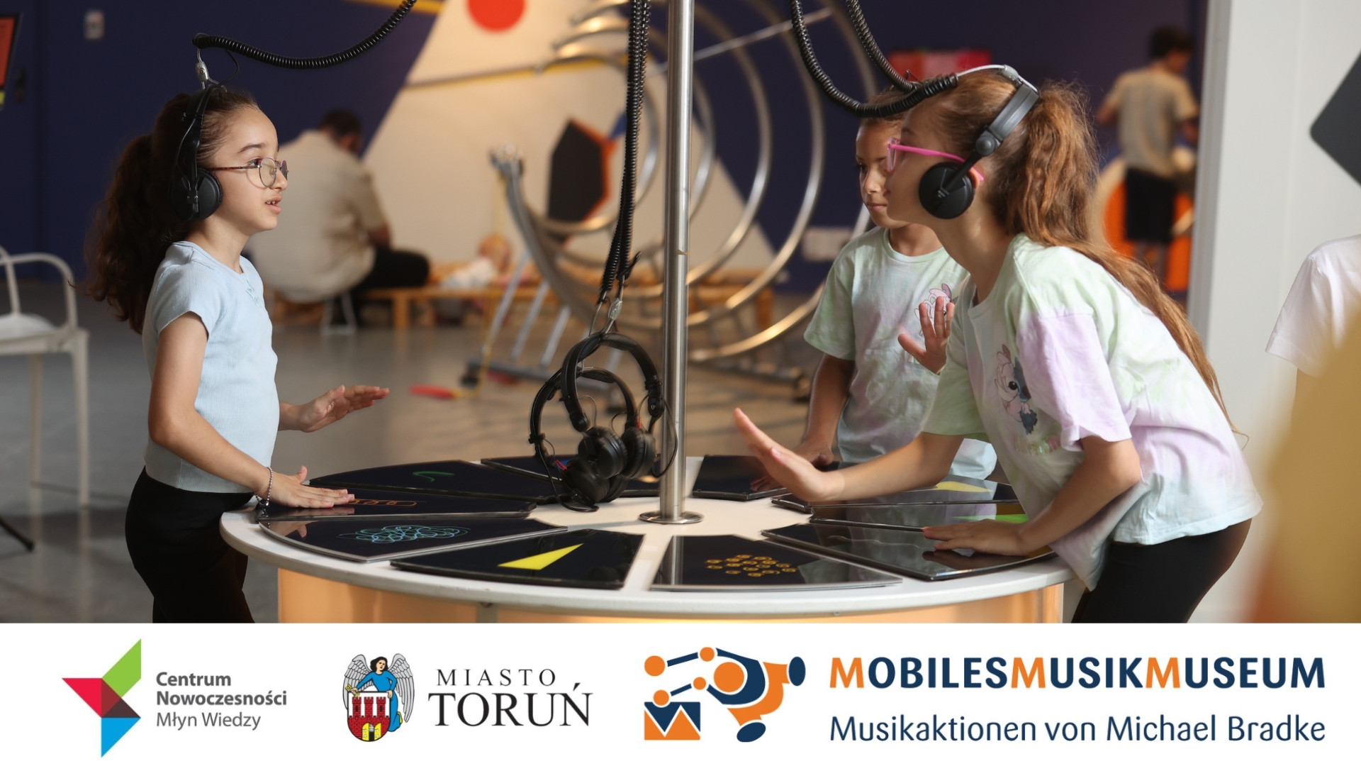 Trzy dziewczynki w słuchawkach na uszach stoją przy eksponacie.