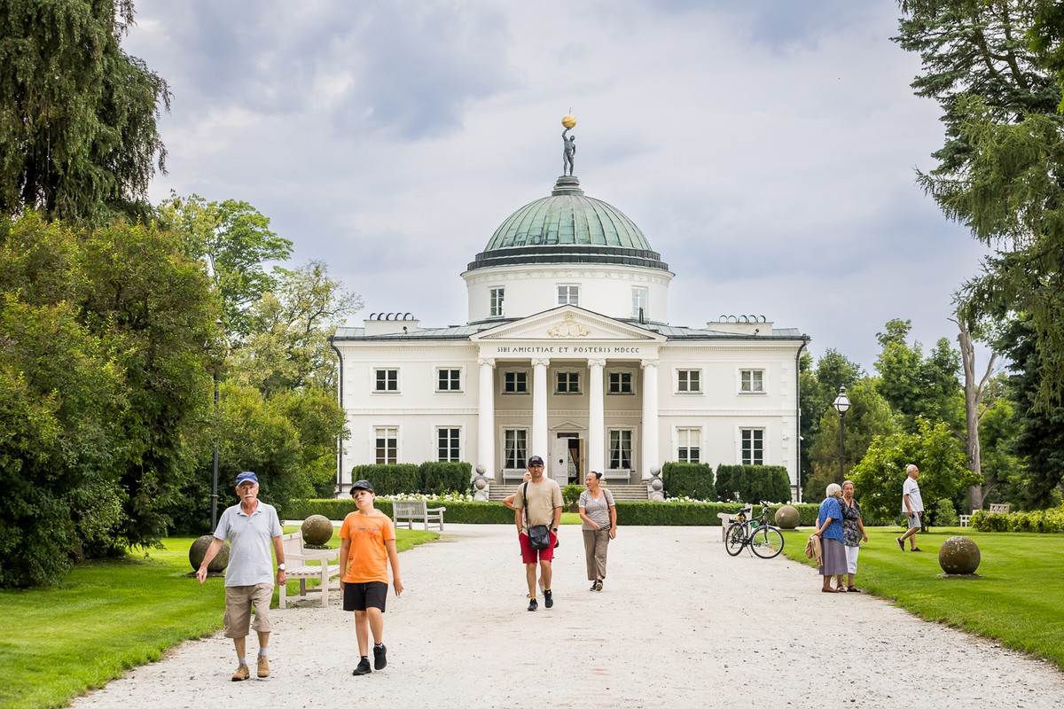 Biały zabytkowy budynek z zieloną kopułą wśród drzew.