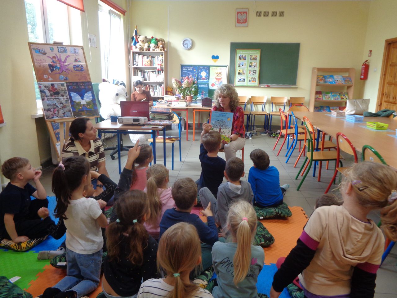 Kobieta prezentuje książkę dzieciom siedzącym na matach.