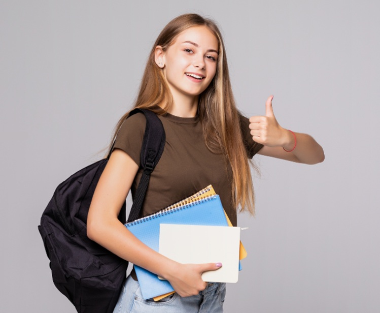 Uśmiechnięta dziewczyna z plecakiem i notatnikami unosi kciuk do góry.