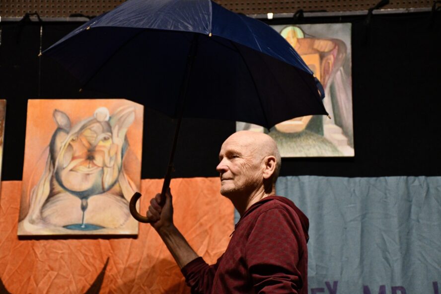 Aktor na scenie trzyma otwarty parasol. W tle obrazy abstrakcyjne.