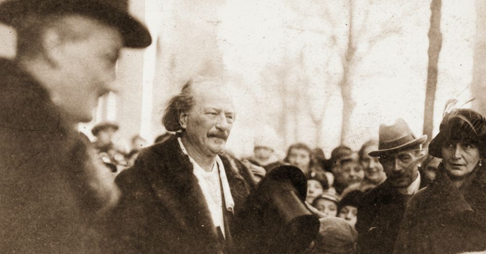 Ignacy Paderewski trzyma cylinder w dłoni. Wokół niego skupieni są dorośli w kapeluszach i dzieci.