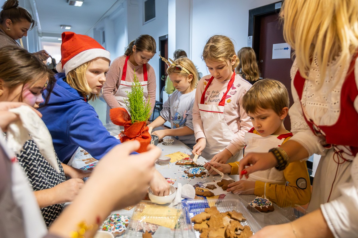 Dzieci w fartuszkach kuchennych dekorują świąteczne pierniczki stojąc wokół stołu z choineczką.