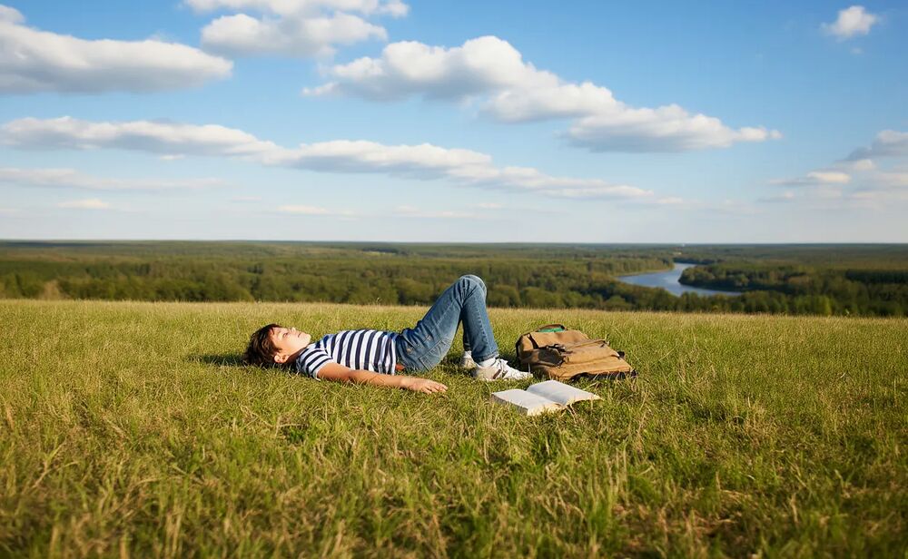 Chłopiec leży na trawie, obok książka i plecak, w tle rzeka i las.