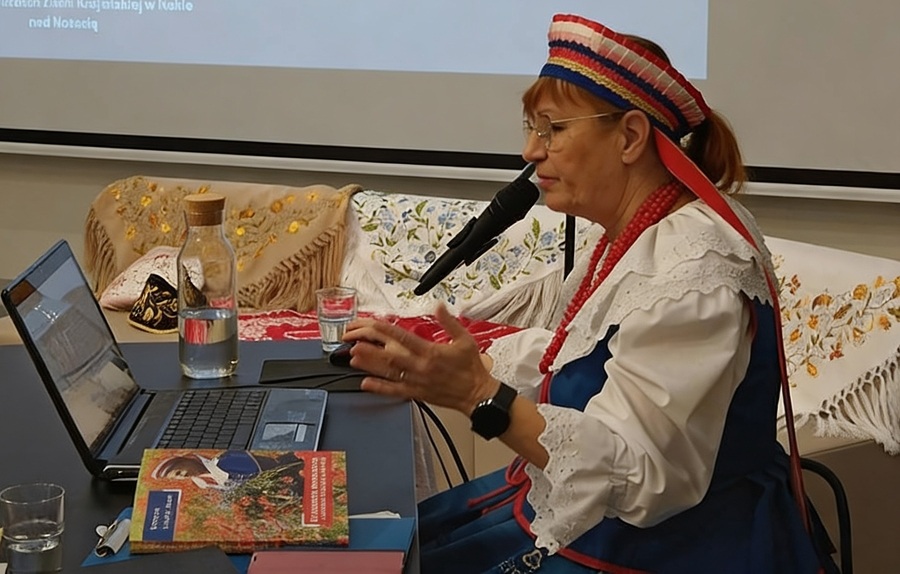 Kobieta w stroju regionalnym przy laptopie podczas wykładu. Na dużym ekranie wyświetlana jest prezentacja.