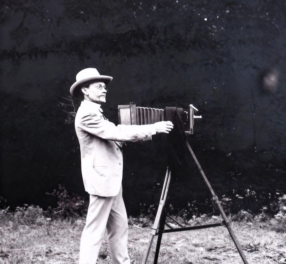 Mężczyzna w garniturze, kapeluszu, w binoklach stoi przy mieszkowym aparacie fotograficznym.