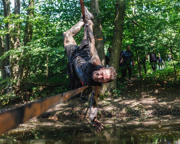 Ubłocony mężczyzna w sportowym stroju pokonuje terenową przeszkodę nad rzeczką.