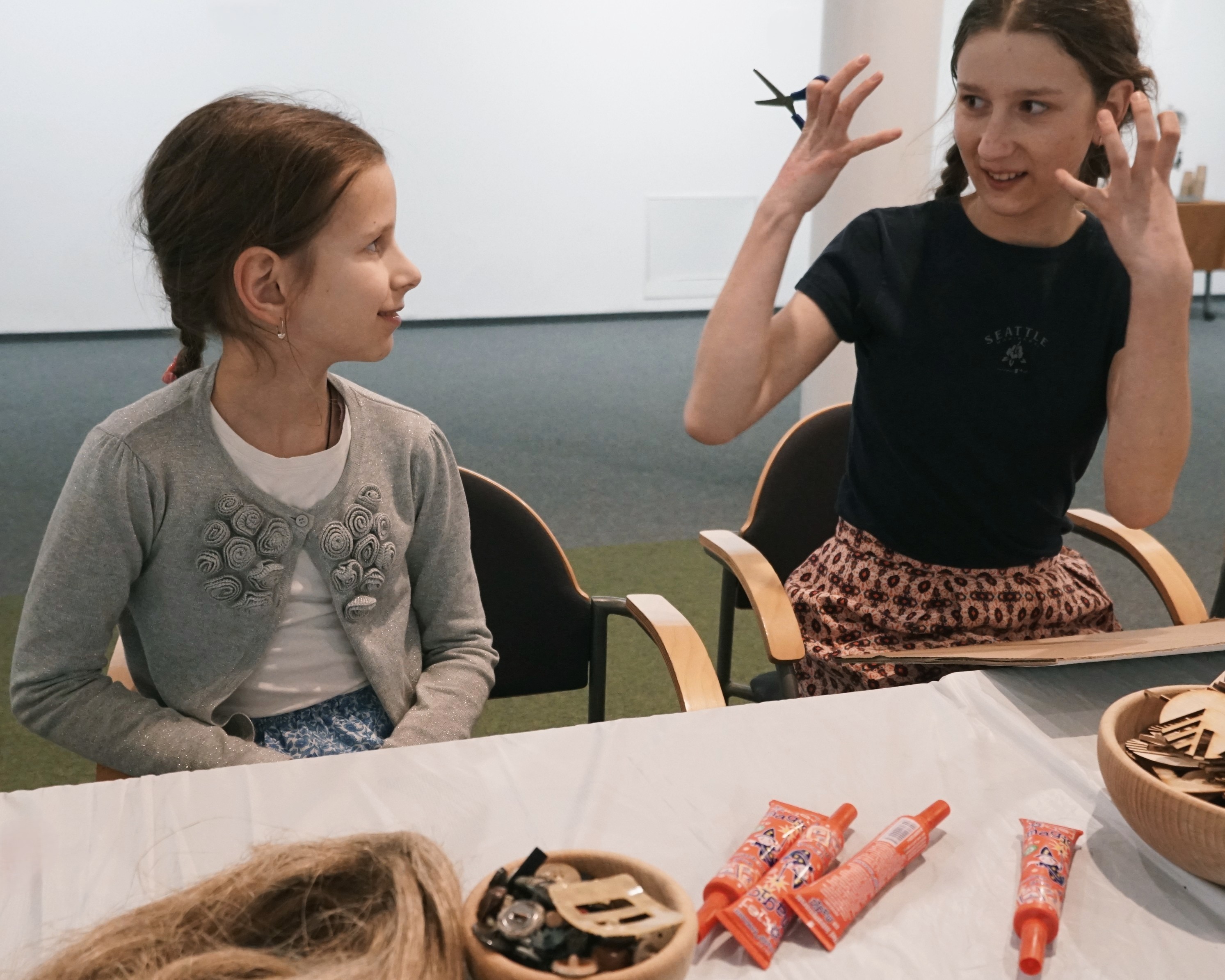 Uśmiechnięte dziewczynka i młoda kobieta z uniesionymi rękami siedzą przy stole z przyborami plastycznymi.