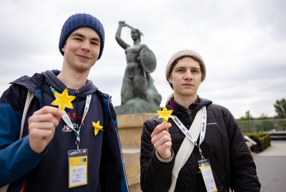 Dwaj nastolatkowie z identyfikatorami trzymają papierowe żonkile, w tle pomnik Syrenki Warszawskiej. 