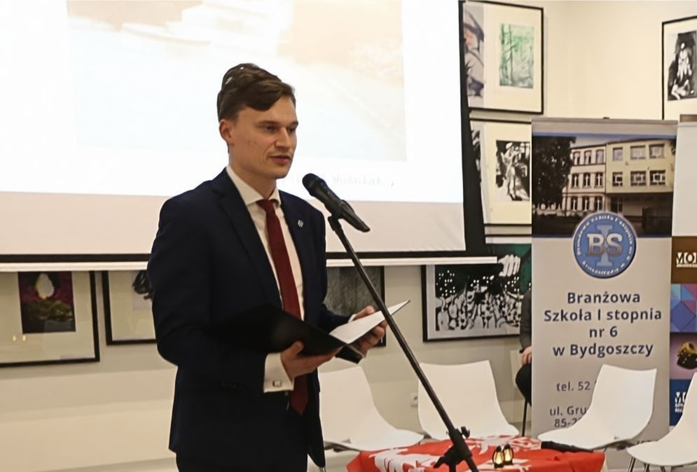 Młody mężczyzna przed mikrofonem. W tle duży ekran i obrazy.