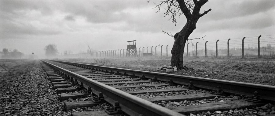 Czarno-biały widok na tory i ogrodzenie z drutem kolczastym oraz wieżyczkę strażniczą obozu.