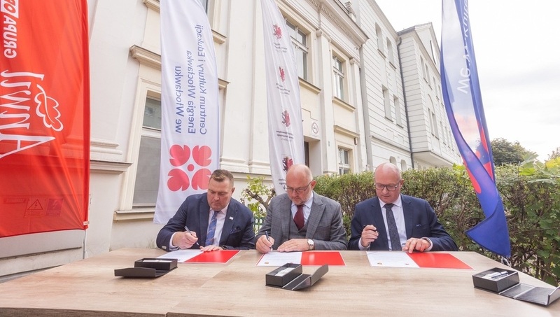 Trzej oficjele podpisują dokumenty przed budynkiem.