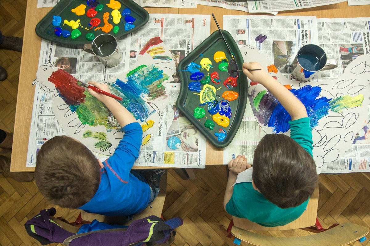 Zdjęcie robione z góry, dwaj chłopcy malują pędzlami, obok palety z kolorowymi farbami.