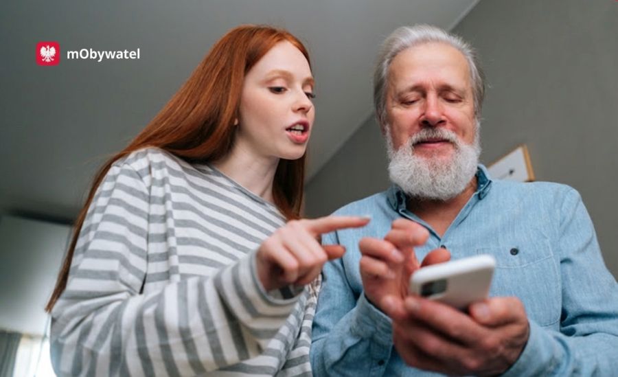 Dziewczyna i starszy mężczyzna spoglądają na ekran smartfona. U góry logo mObywatela.