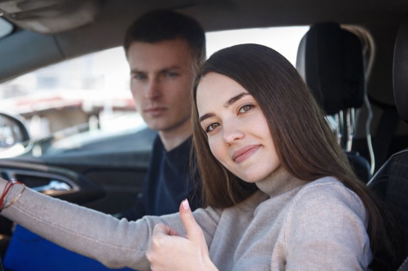 Dwoje młodych ludzi w samochodzie. Kobieta za kierownicą unosi kciuk w górę.