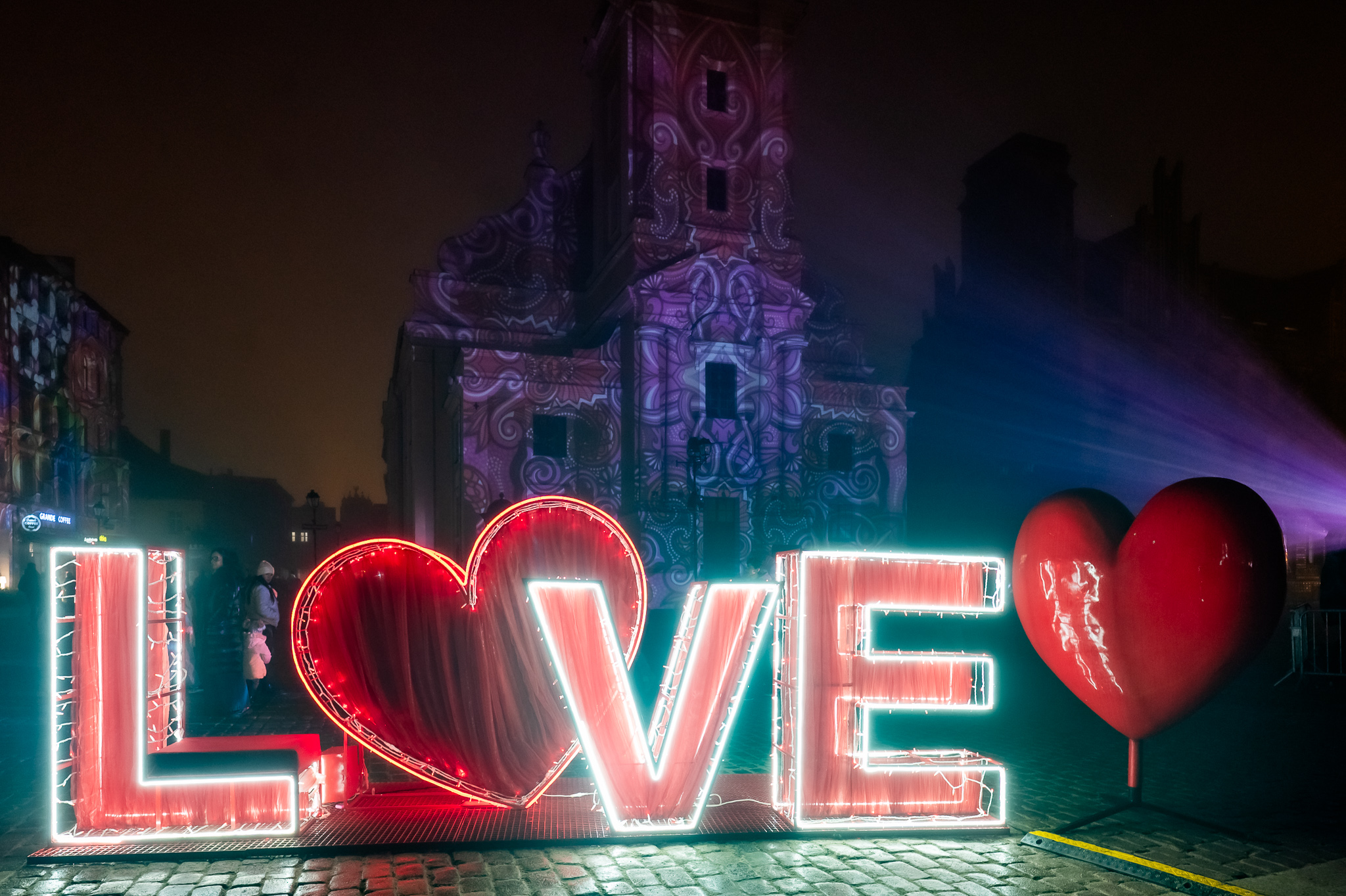 Miasto wieczorem, świecący napis LOVE z sercem na tle budynku z mappingiem.