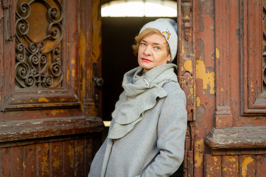 Kobieta opiera się o stare drzwi z metalowymi zdobieniami i odpadającą farbą, w tle klatka kamienicy.