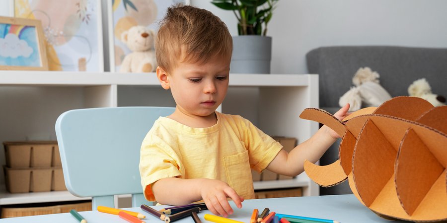 Mały chłopiec siedzi przy stoliku z kredkami i figurą zrobioną z tektury. 