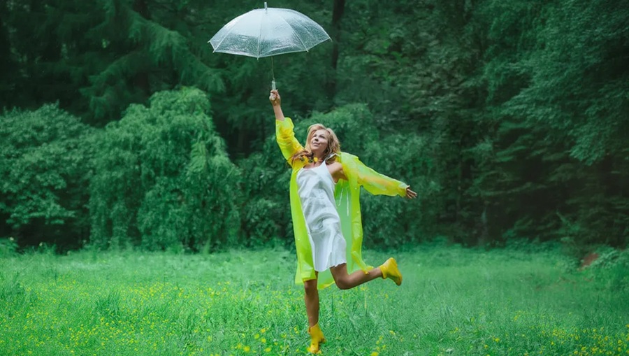 Kobieta w płaszczu przeciwdeszczowym podskakuje i unosi wysoko parasol. W tle łąka i las.
