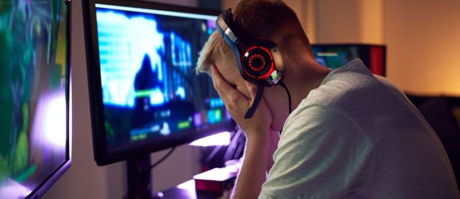 Młody mężczyzna w słuchawkach opiera głowę na dłoniach, kryjąc w nich twarz. Przed nim trzy monitory.