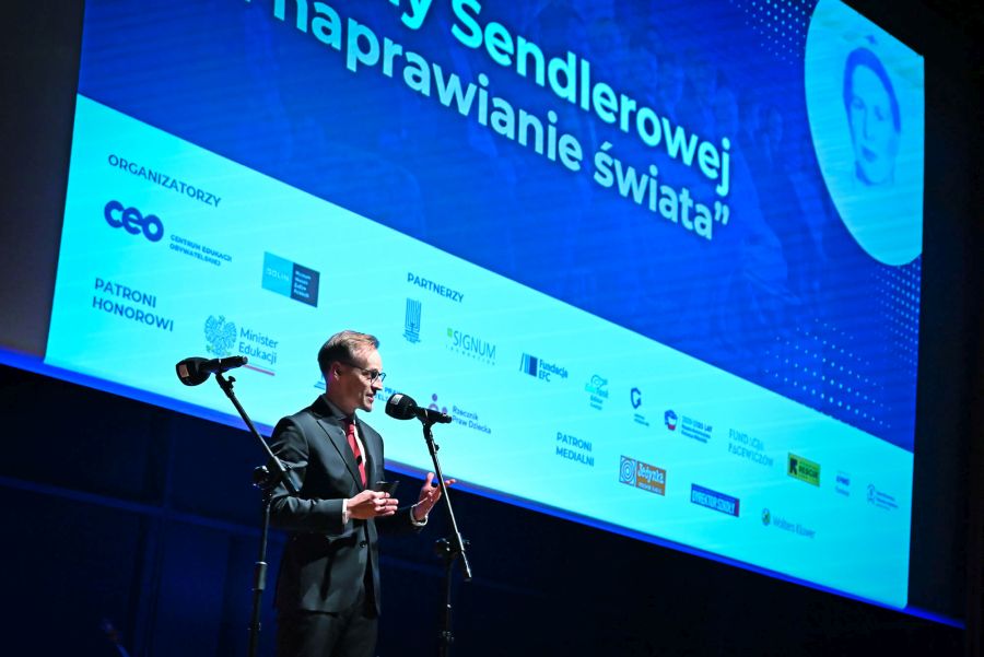 Mężczyzna na scenie przy mikrofonie. Za nim ekran z napisem: Nagroda „Za naprawianie świata” im. Ireny Sendlerowej.