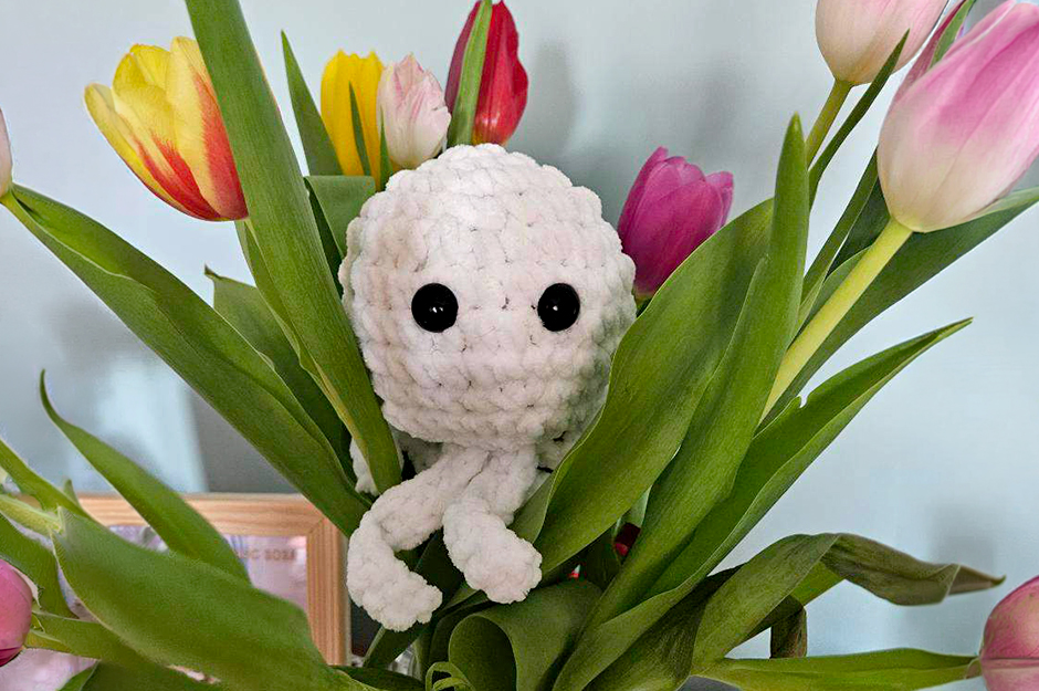 Stworek zrobiony na szydełku wśród kwiatów.