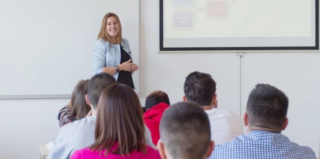 Słuchacze i kobieta prowadząca zajęcia przy ekranie z prezentacją.