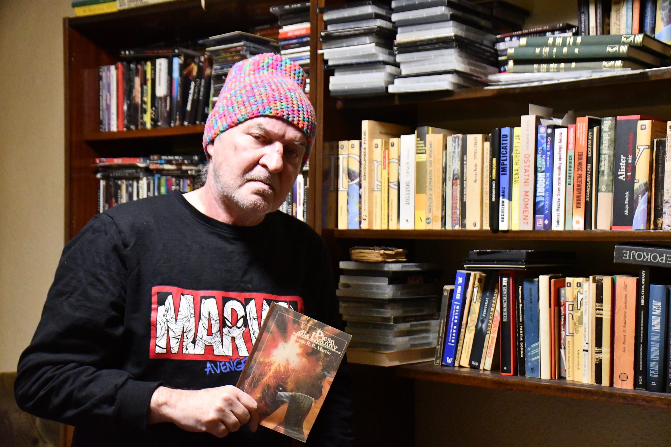 Mężczyzna w kolorowej czapce stoi przy regale z książkami.