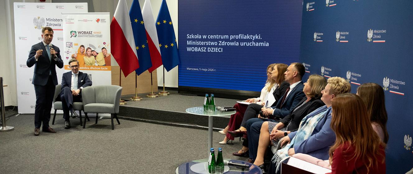Urzędnicy ministerstwa siedzą w rzędzie. Za nimi ekran z prezentacją programu badań profilaktycznych dzieci i młodzieży. 