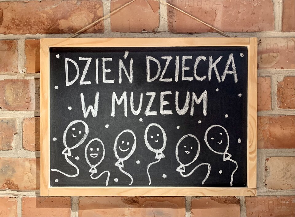 Na ścianie z cegieł powieszona jest tablica z napisem białą kredą Dzień Dziecka w muzeum.