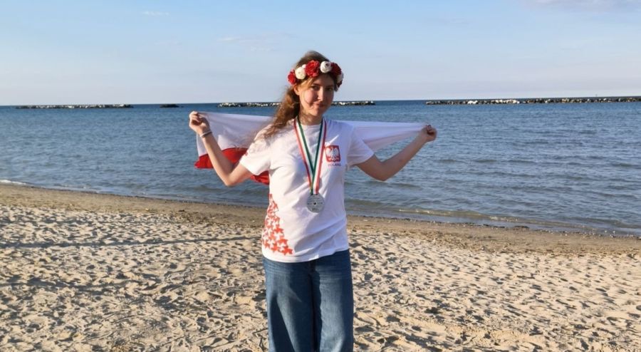 Na plaży stoi dziewczyna z medalem na szyi, wiankiem w barwach narodowych i flagą biało-czerwoną.