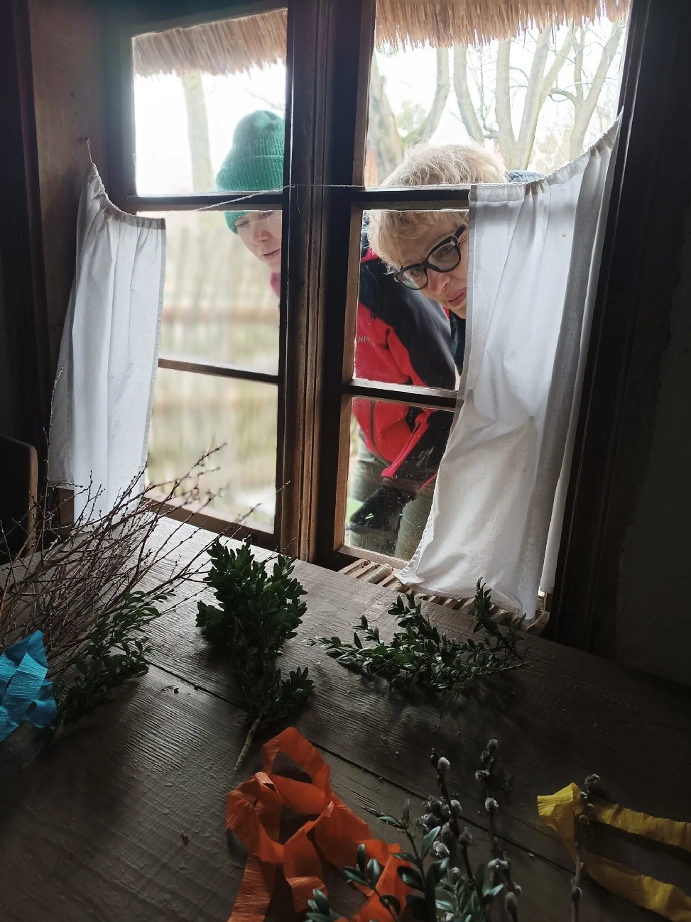 Fragment drewnianego stołu, leżą na nim palmy, drewniane okno przez które zaglądają dwie osoby.