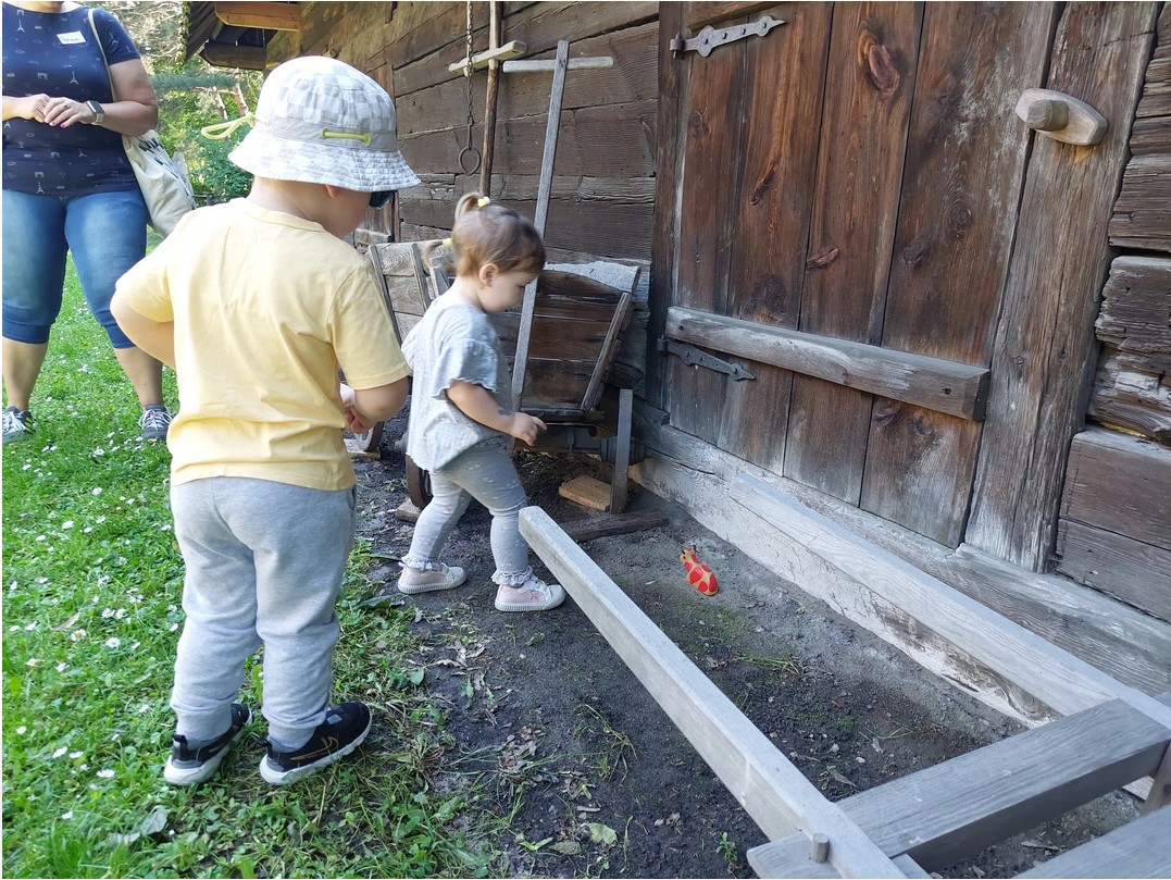Dwójka małych dzieci spogląda w kierunku drewnianej figurki. W tle ściana budynku z drewna.