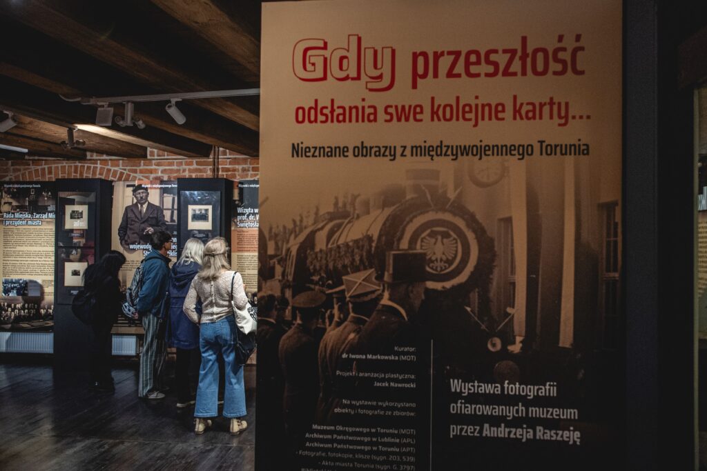 Sala w muzeum, na pierwszym planie plakat promujący wystawę, w tle trzy osoby oglądają wystawę.
