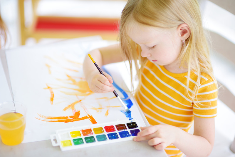 Mała dziewczynka o jasnych włosach maluje na papierze kolorowymi farbami