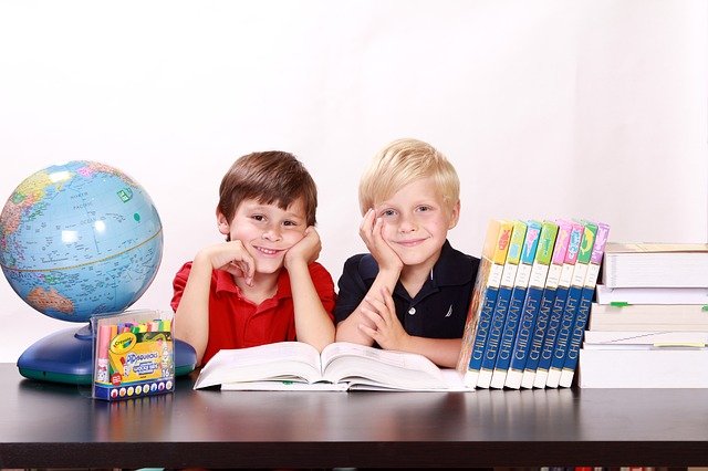 Dwaj uśmiechnięci chłopcy siedzą nad otwartą książką leżącą na stole, obok stoją książki, globus i kolorowe pisaki