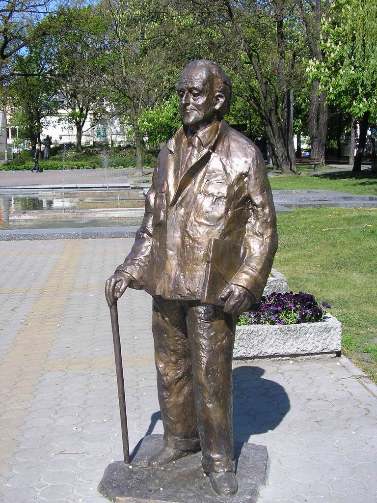 Pomnik z brązu Andrzeja Szwalbego z laską na tle zieleni miejskiej.
