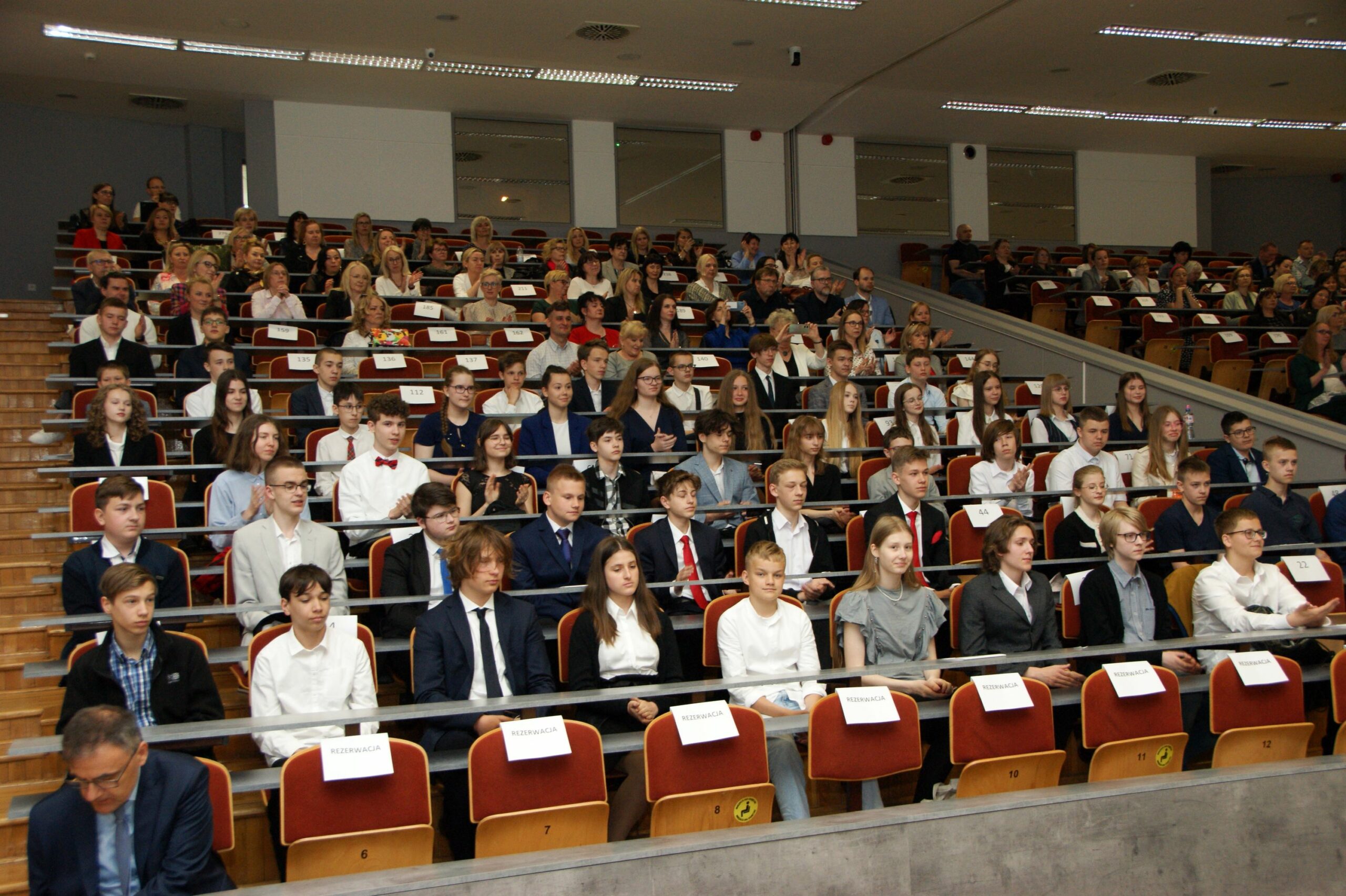 Młodzież i nauczyciele w auli uczelni.