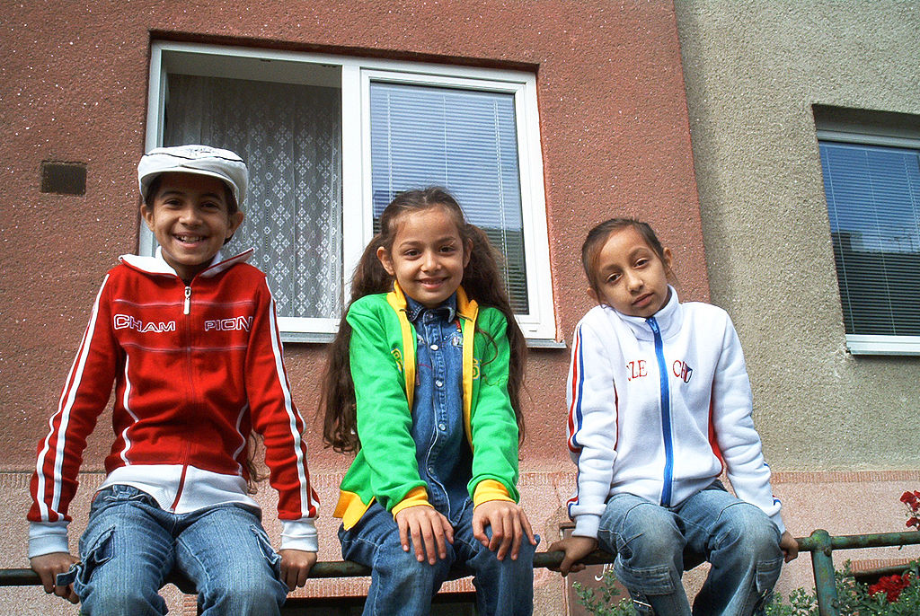 Troje romskich dzieci siedzących przed budynkiem.