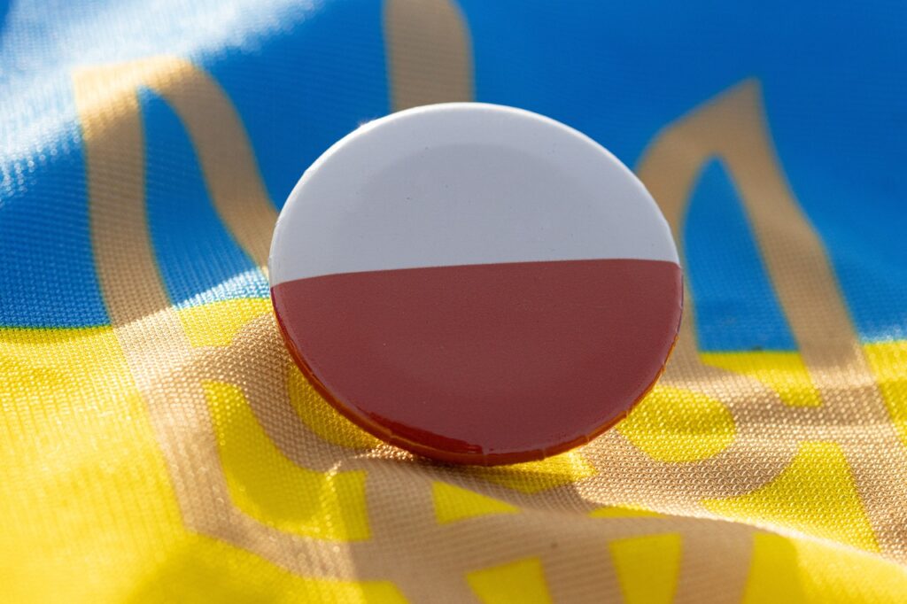 Okrągła przypinka w barwach biało-czerwonych na tle ukraińskiej flagi z trójzębem.
