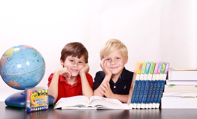 Dwaj uśmiechnięci chłopcy siedzą nad otwartą książką leżącą na stole, obok stoją książki, globus i kolorowe pisaki