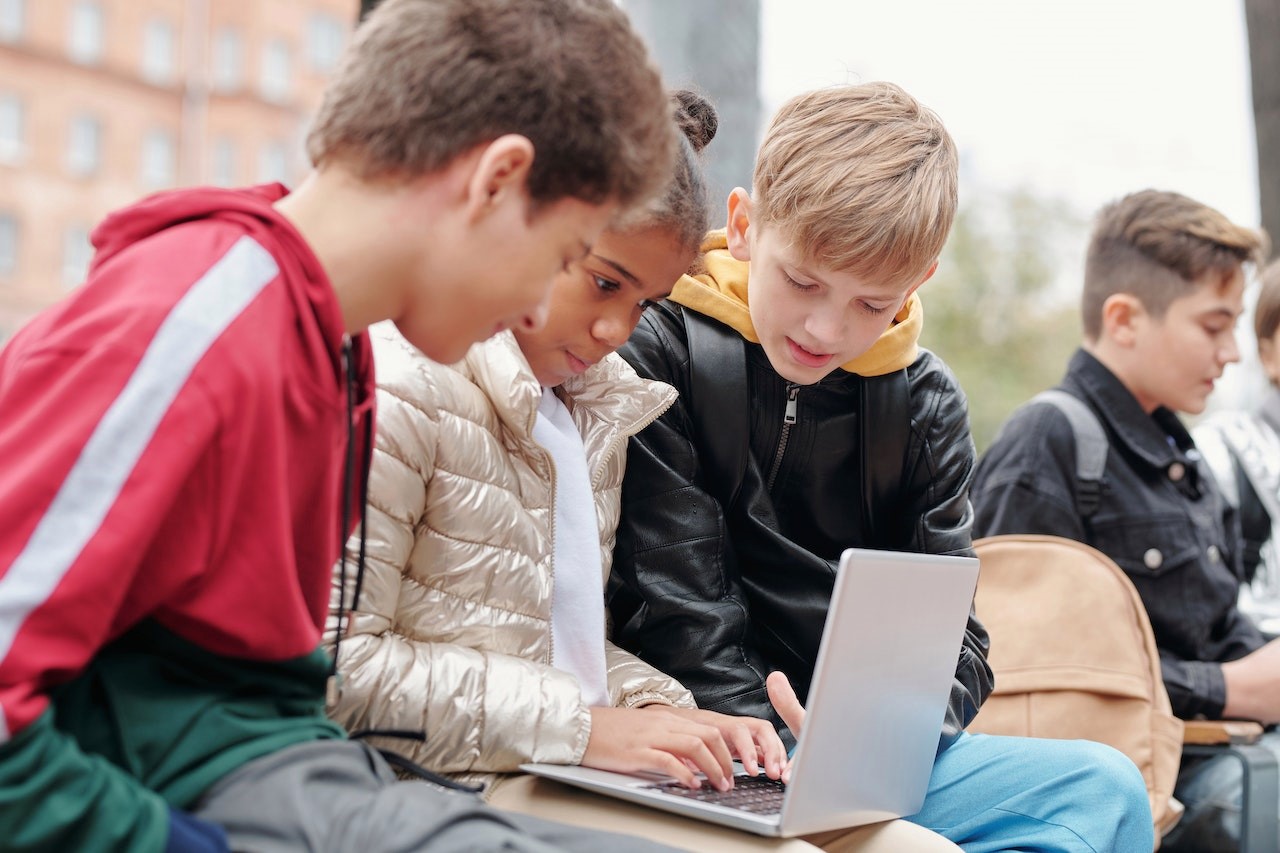 Uczniowie siedzą przed budynkiem i patrzą w laptop, który trzyma na kolanach dziewczynka.