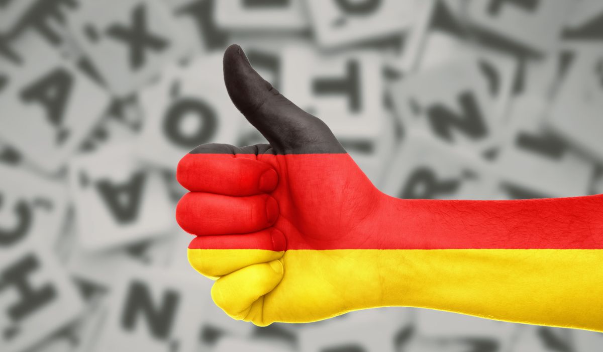 Kciuk uniesiony w górę na dłoni w kolorach niemieckiej flagi. W tle rozsypane litery.