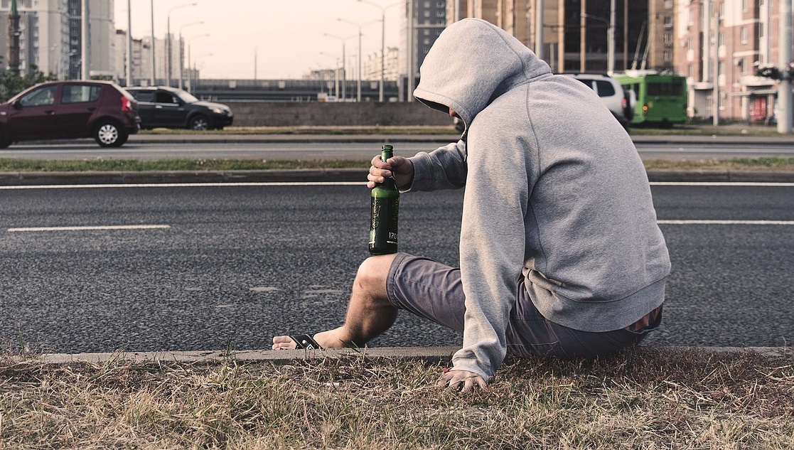 Mężczyzna z kapturem na głowie siedzi na krawężniku drogi z butelką alkoholu.