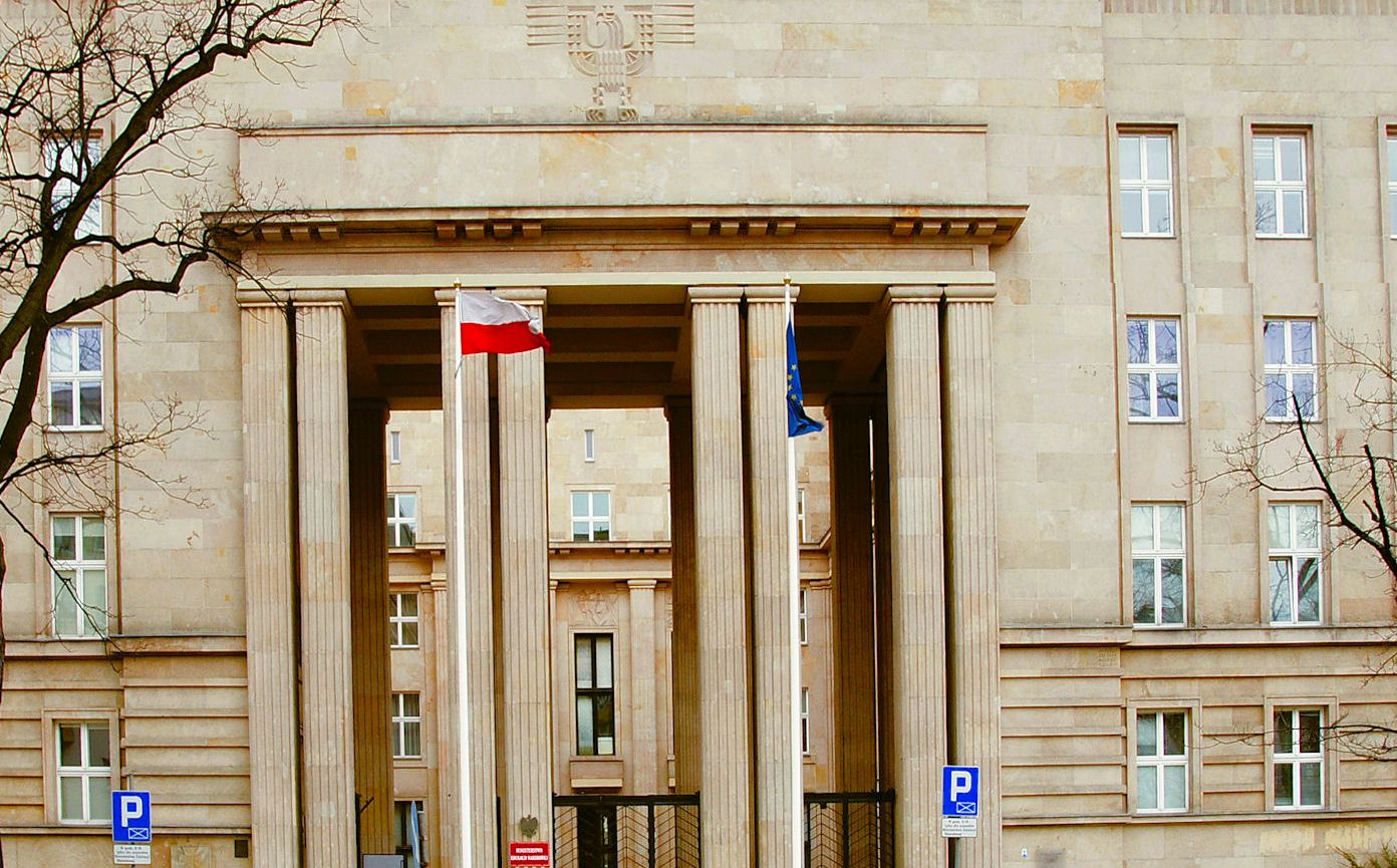Fragment fasady budynku w stylu neoklasycystycznym. Przed wejściem flaga narodowa i Unii Europejskiej na masztach.
