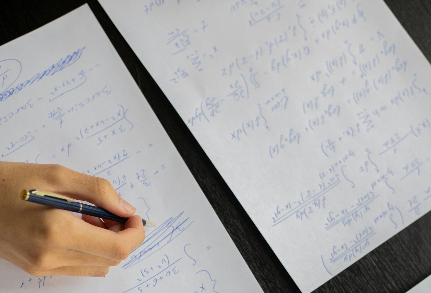 Dłoń z długopisem na kartkach z zapisami matematycznymi.