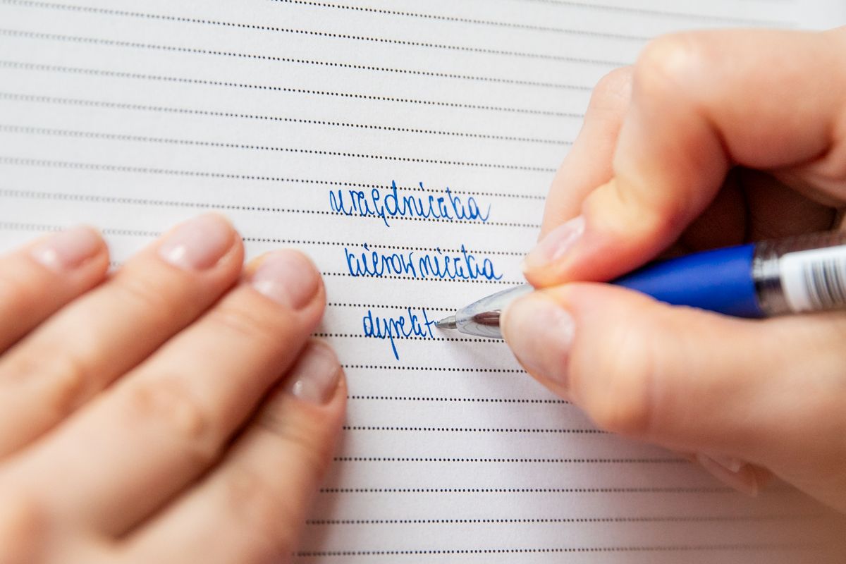 Dłoń z długopisem na liniowanym papierze zapisuje feminatywy: urzędniczka, kierowniczka, dyrektorka.